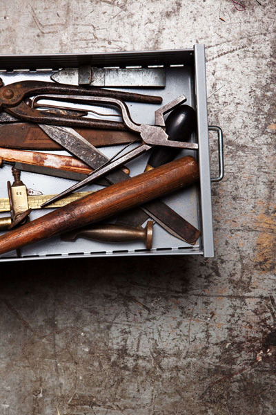 Tiroir métallique ouvert révélant des outils anciens de maroquinerie : pinces, cisailles, alênes et maillet en bois, sur une surface d'atelier vieillie